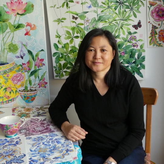 Photo og Gabby sitting at a small table covered in her colourful artworks, the work is delicate and has things like Chinese decorative pots, lotus flowers, green leaves and peonies. Gabby has a black top on, she has black hair and brown skin and smiles warmly. She sits on a wooden chair.